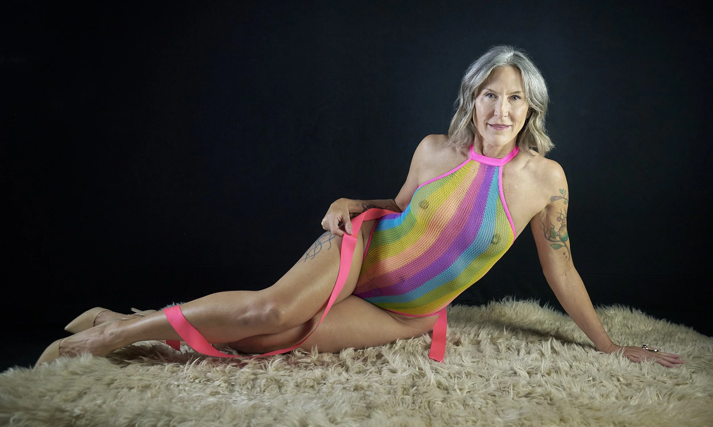 A woman with gray hair posed on a furry rug against a black background, wearing a colorful, rainbow-striped mesh bodysuit and beige high heels.