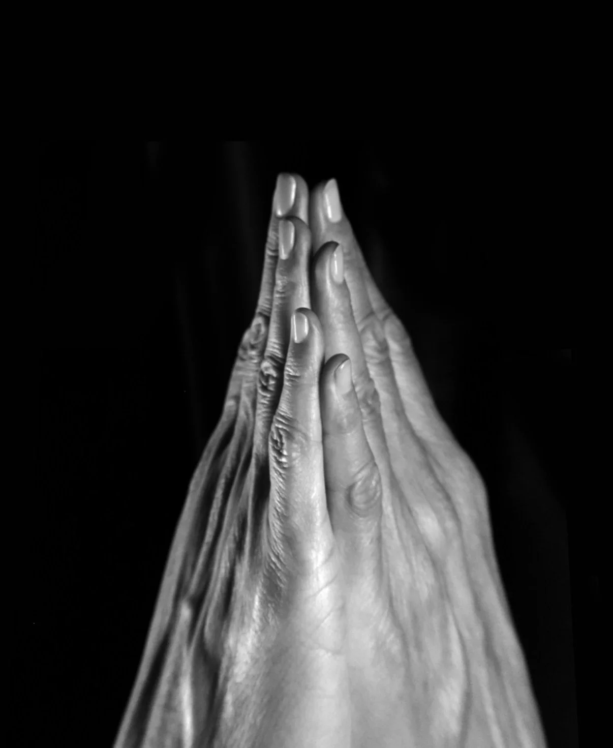 Black and white photograph of multiple hands pressed together in prayer or meditation, with fingers pointing upward, on a dark background.