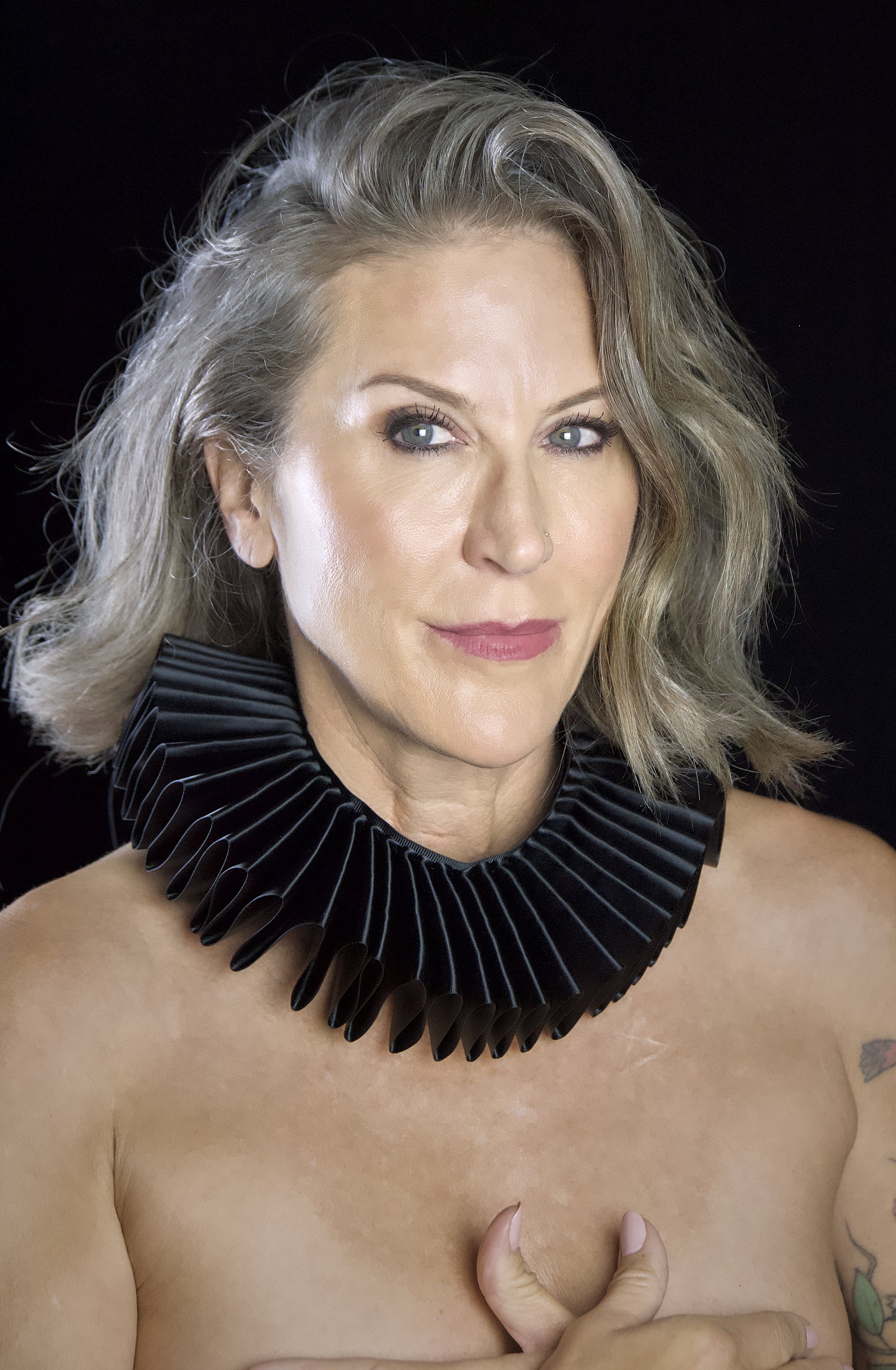 A woman with blonde hair and blue eyes wearing a large black pleated collar, posing against a black background.