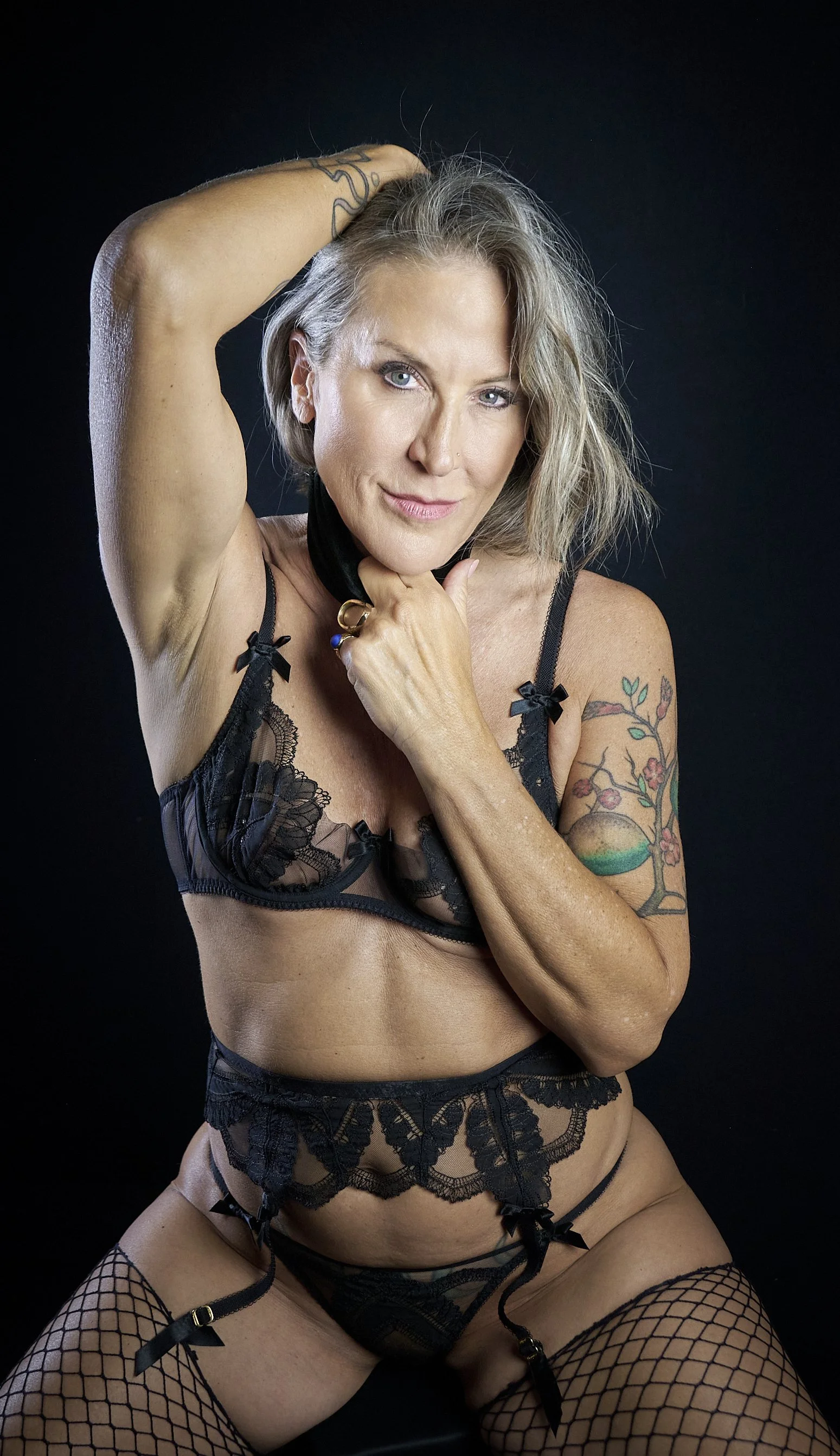 A woman with blonde hair and tattoos wearing black lace lingerie and fishnet stockings posing against a black background.