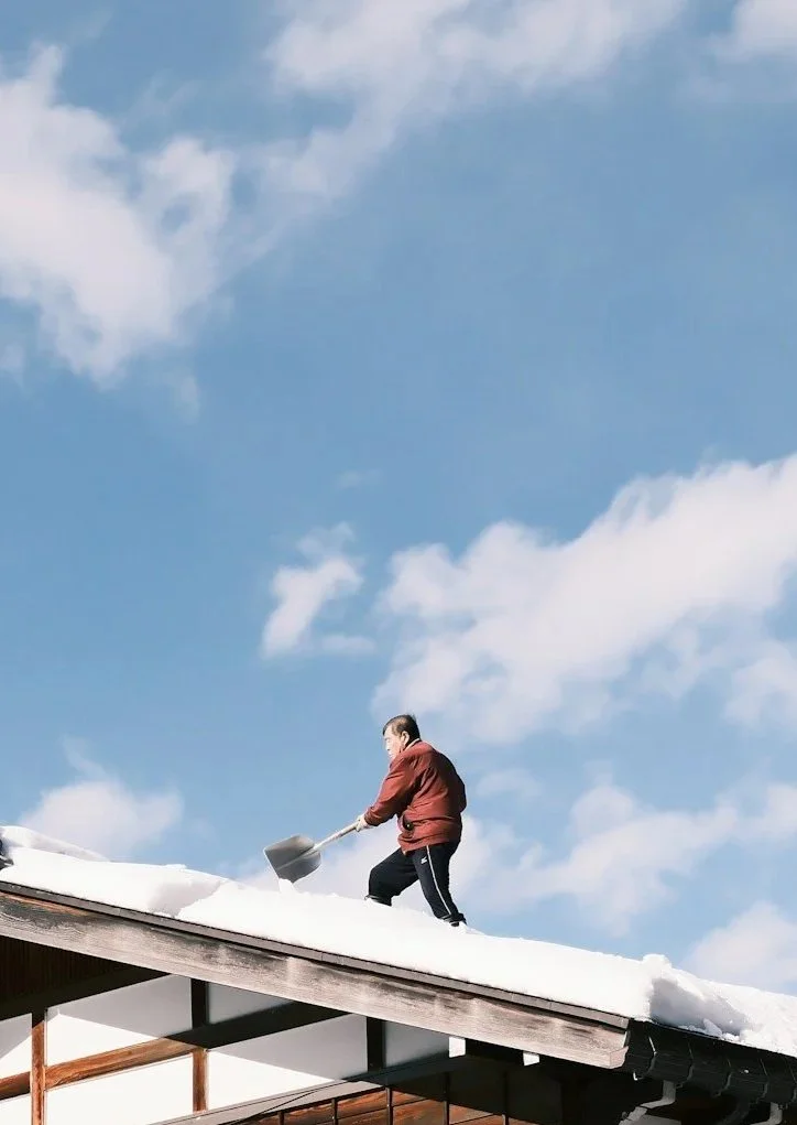 Person shoveling snow off the roof of a house during the daytime with a blue sky and clouds in the background.