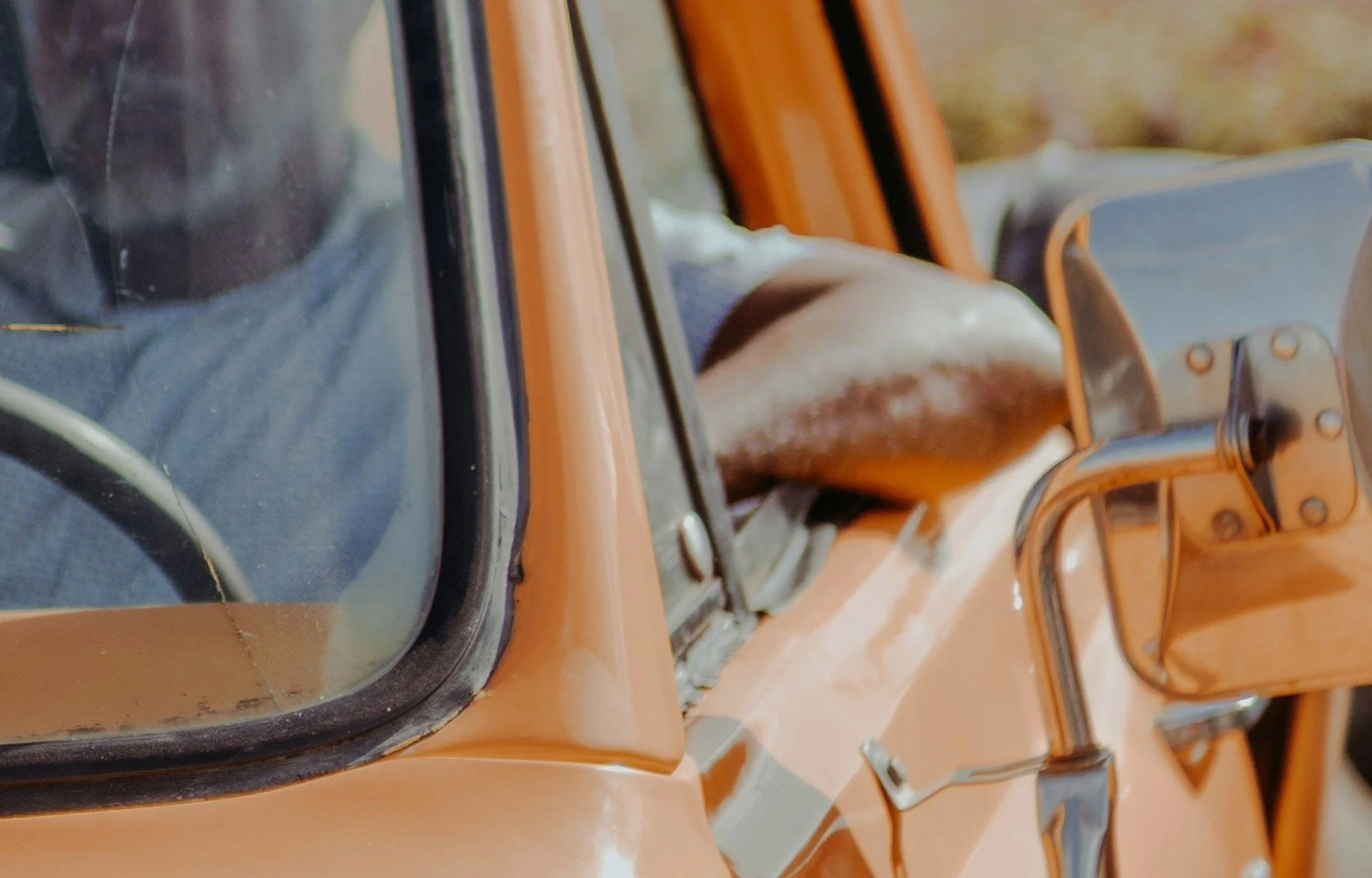 Partial view of a vintage orange car, focusing on the side mirror, window, and part of the door, with clear outdoor background.