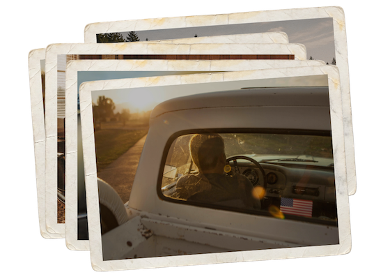 View from behind the windshield of a vintage truck during sunset, with a person driving and another passenger sitting beside, on a rural road.