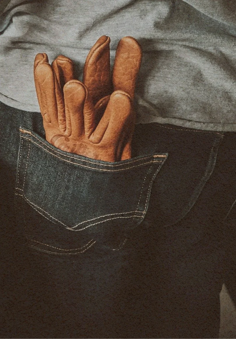 Close-up of a person wearing dark jeans with a back pocket and a gray t-shirt, with one hand in the pocket wearing brown work gloves.