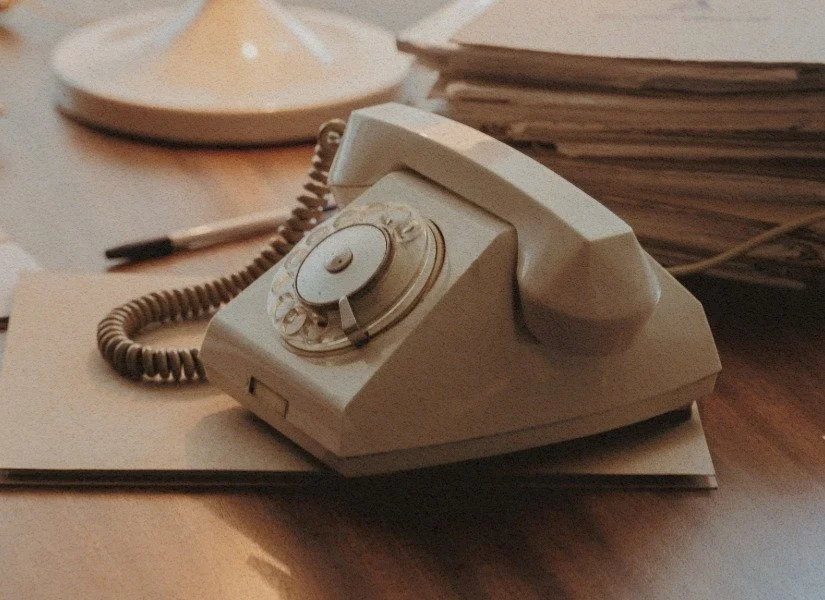 Close-up of a vintage rotary dial telephone on a desk with papers, a pen, and a dish in the background.