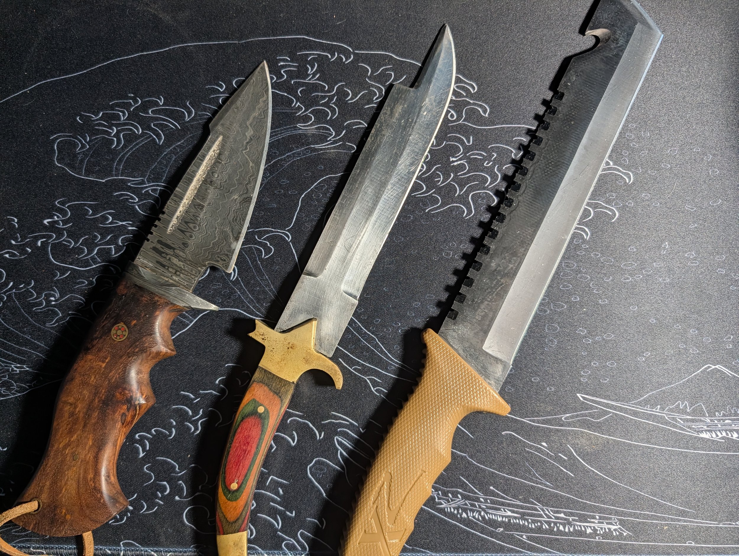 Three survival knives with different handles and blade styles resting on a black surface with a white line drawing of a wave or cloud pattern.