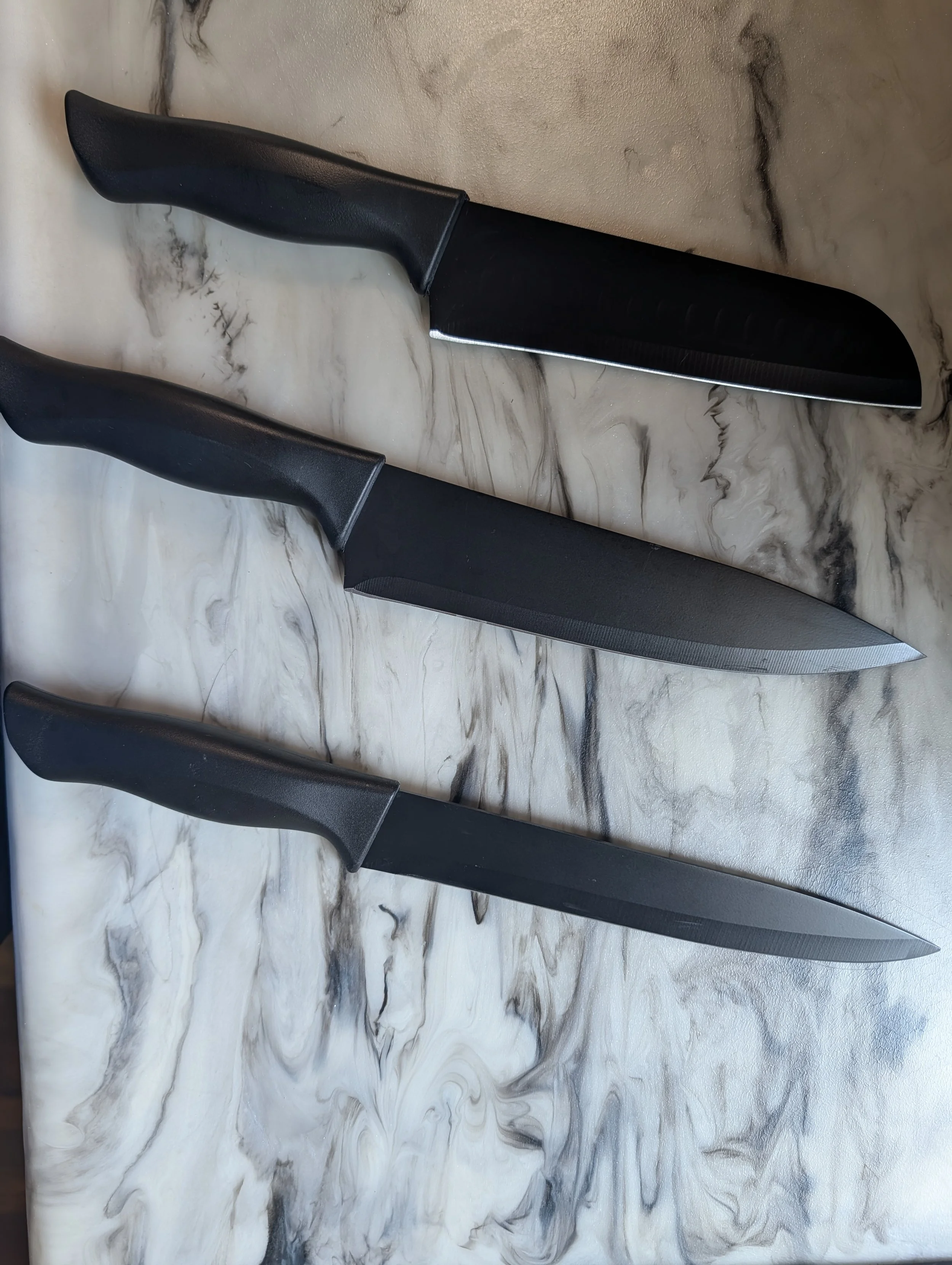 Two kitchen knives with black handles and blades are placed on a marble surface.