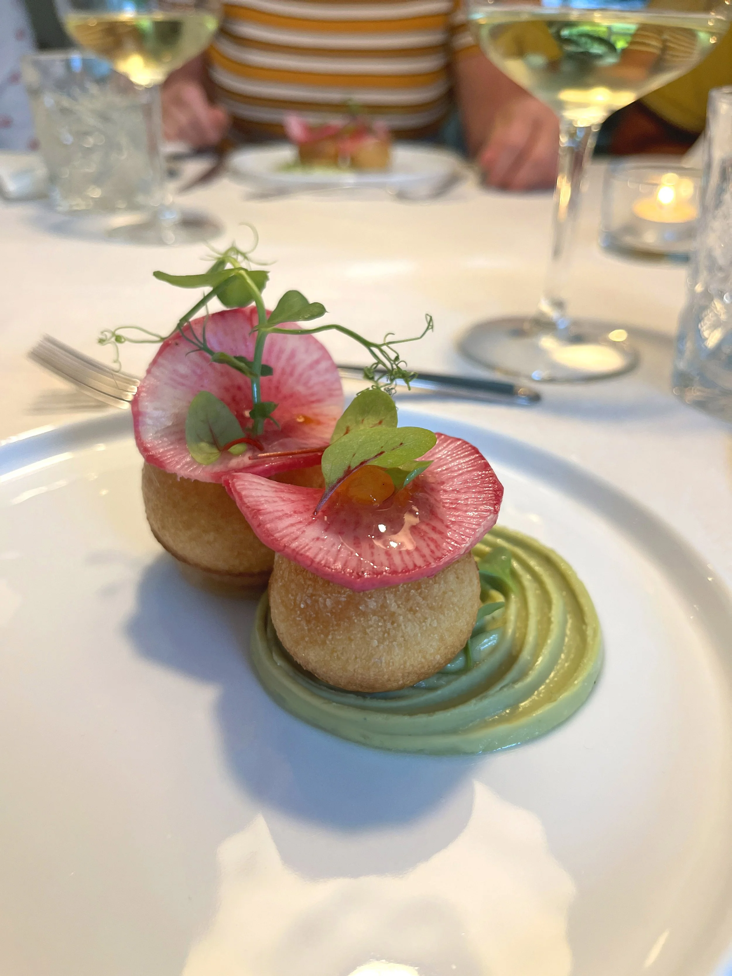 Een amusegerecht met een gefrituurde balletje op een bedje van groene mousseline en decoratie van radijs en microgroenten, geserveerd op een wit bord.