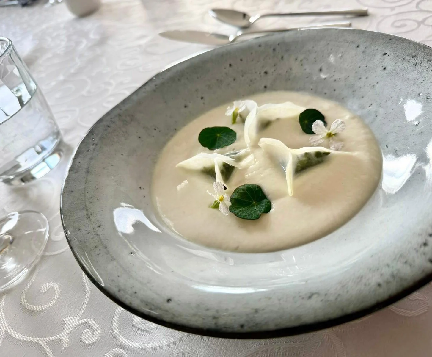 Een witte, romige soep geserveerd in een kom met groene bladeren en witte bloemen als garnering, op een witte tafelkleed.