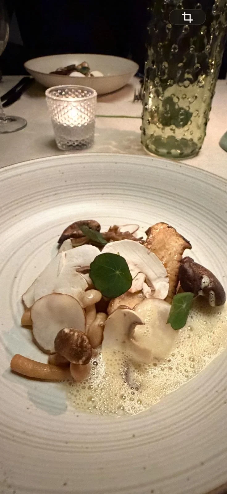Gerechten met verschillende paddenstoelen en groene bladeren op een groot wit bord, op een gedekte tafel met een kaars en een grote groene vaas op de achtergrond.