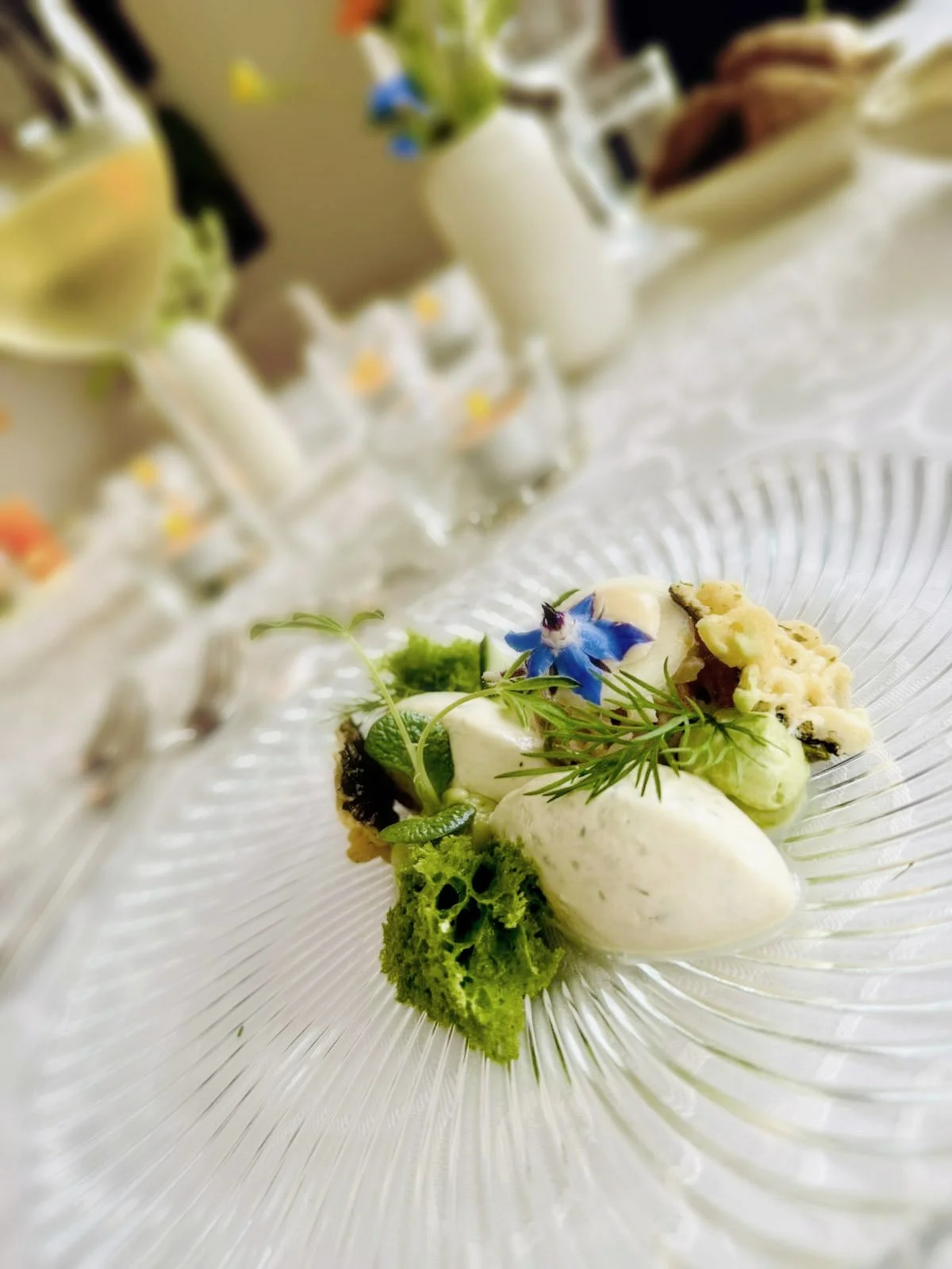 Een gerecht met groene en witte componenten, versierd met een blauwe bloem en takjes, geserveerd op een glazen bord.