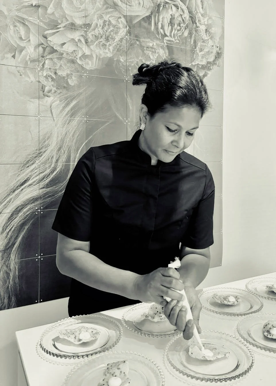 Vrouw die taarten of desserts op kleurrijke glazen borden opsiert, met een bloemendecoratie op de muur op de achtergrond.