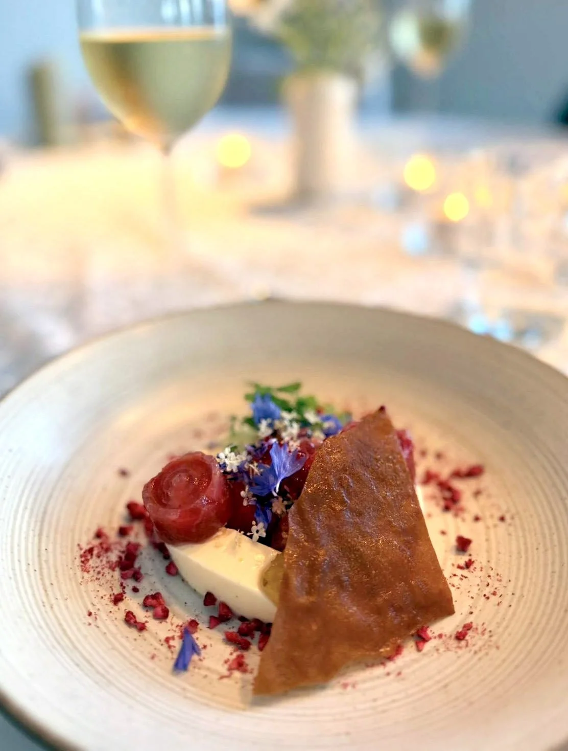 Een dessert met rood fruit, een krokante plaat, blauwe bloemen, en witte saus op een beige bord, met een glas witte wijn op de achtergrond.