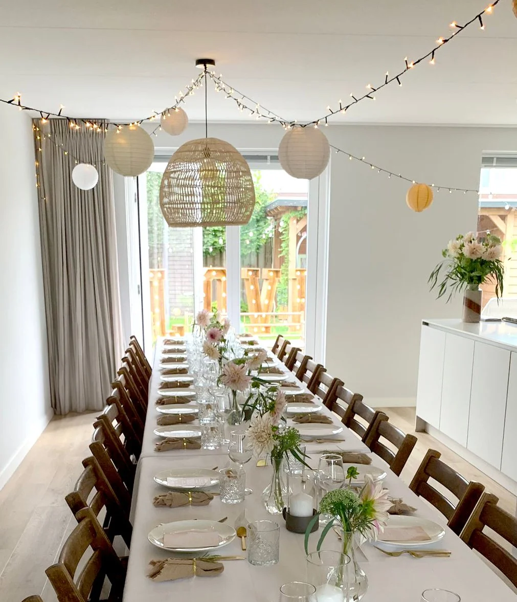 Een feesttafels met bloemen en kaarsen, versierd met paper lanterns en string lights, in een lichte kamer met grote ramen.