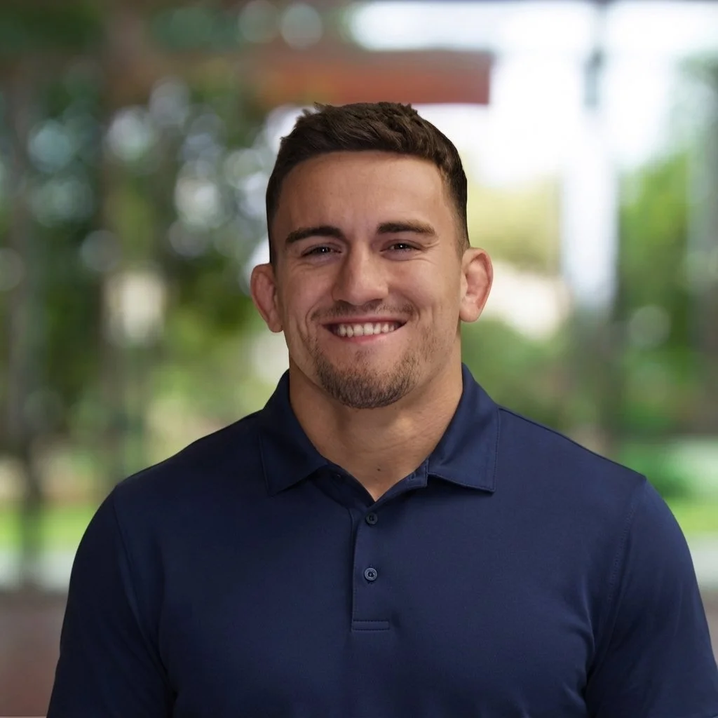 A smiling man with short dark hair and a beard, wearing a navy blue collared shirt, standing outdoors with blurred trees and greenery in the background.