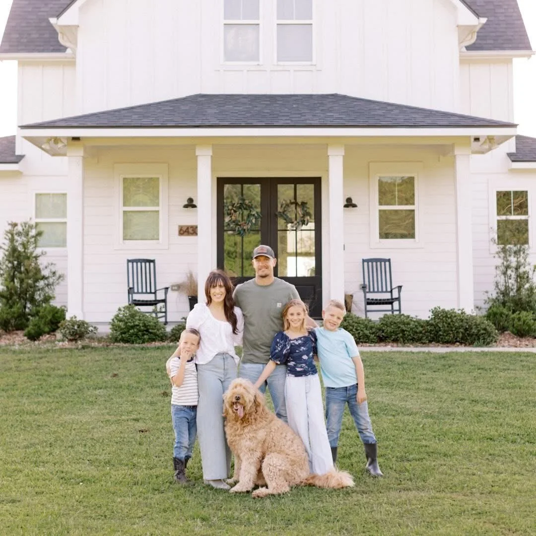 A family of five, including three children, a man, a woman, and a dog, standing in front of a white house on the lawn.
