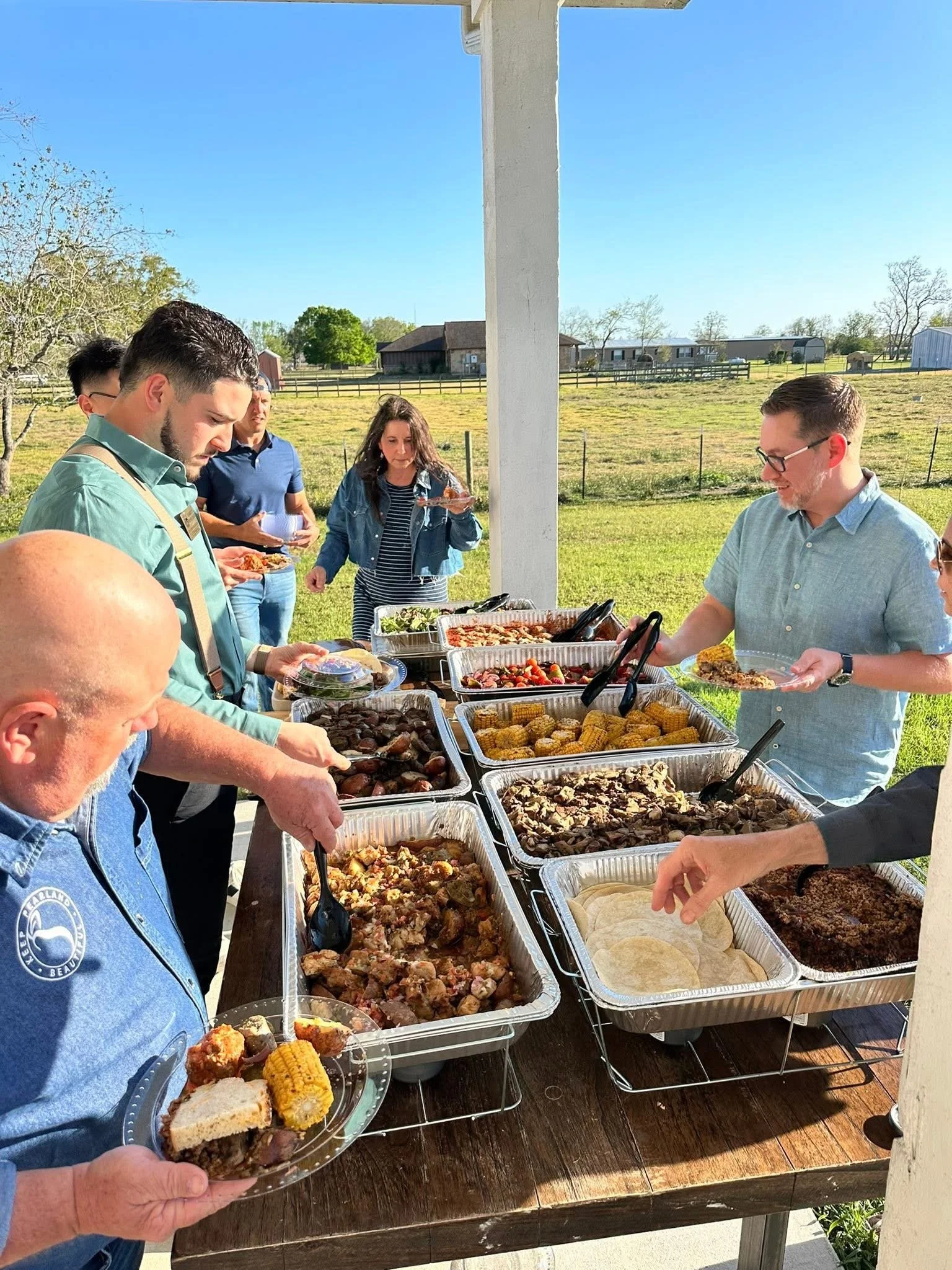 People serving themselves food at an outdoor buffet on a sunny day, with a rural landscape in the background.