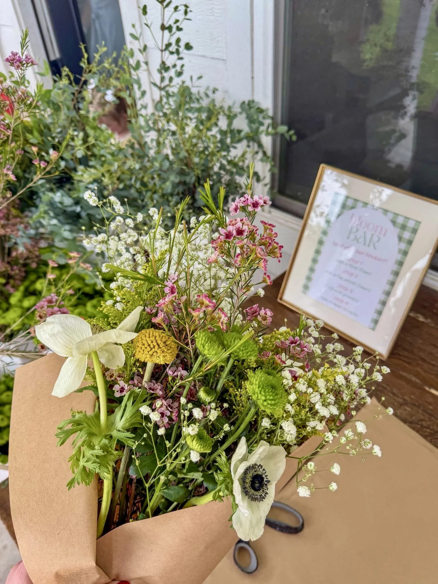 A bouquet of fresh flowers wrapped in brown paper with a framed sign in the background that reads "Bloom & Beir".