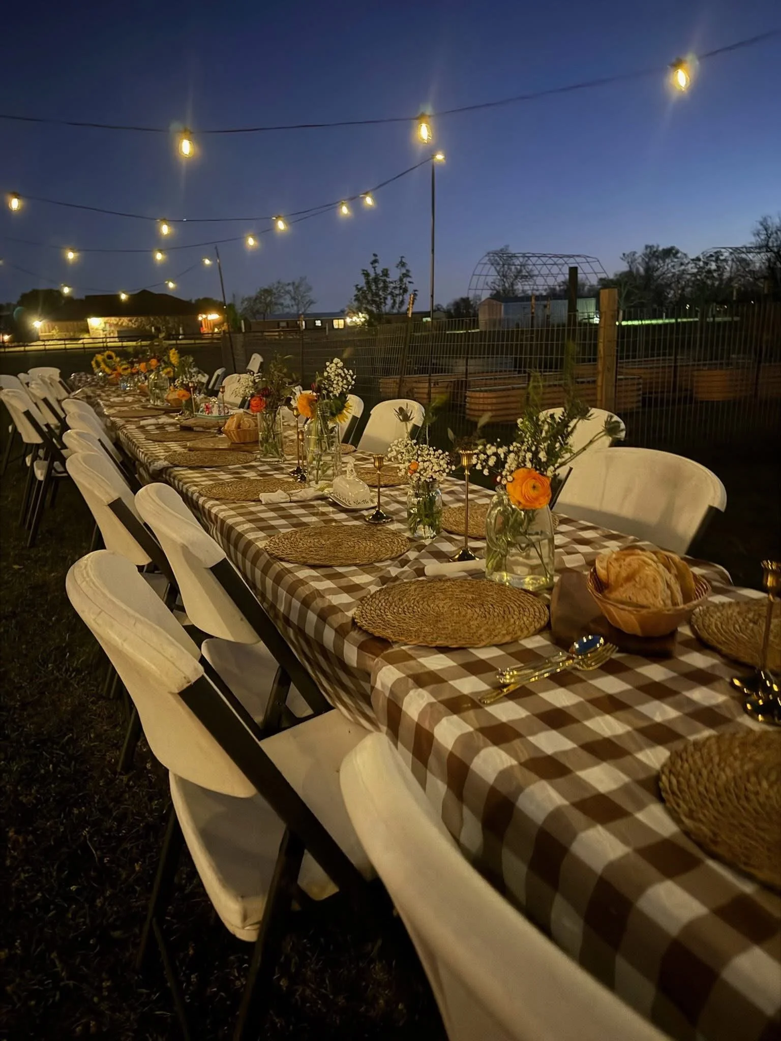 Long outdoor dining table with checkered tablecloth, decorated with flower arrangements, candles, and bread, set for an evening gathering under string lights.