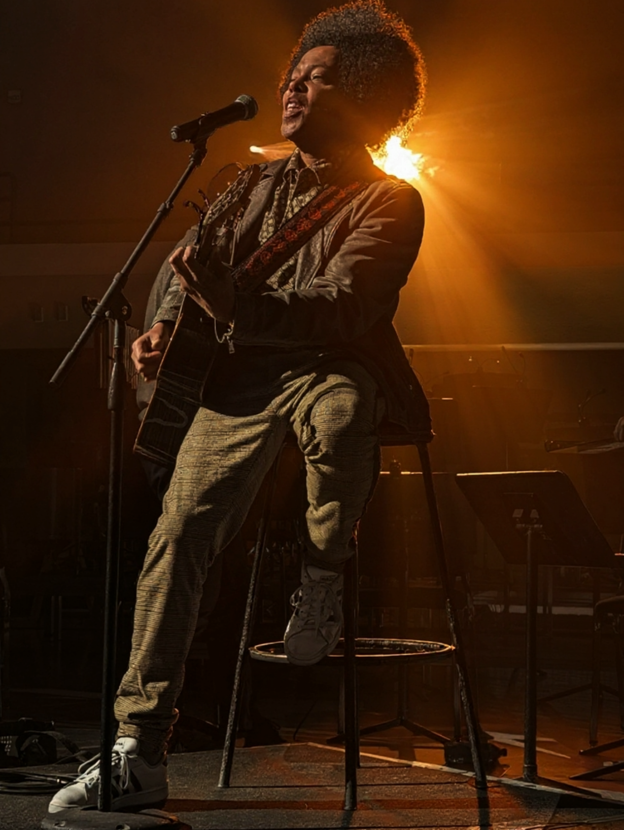 A man singing and playing an acoustic guitar on stage, illuminated by warm backlighting, with one foot on a stool.