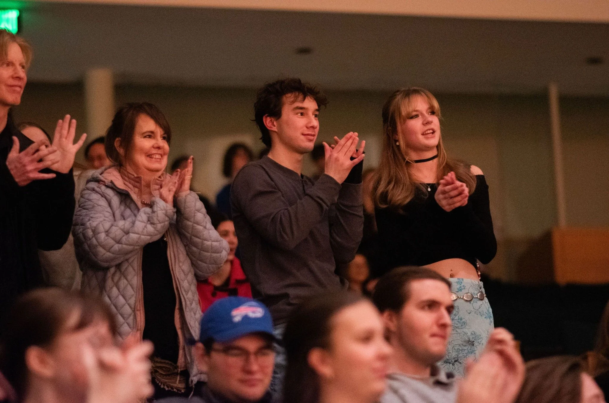 Audience members standing and clapping at an indoor event, with a diverse group of people including men and women of various ages.