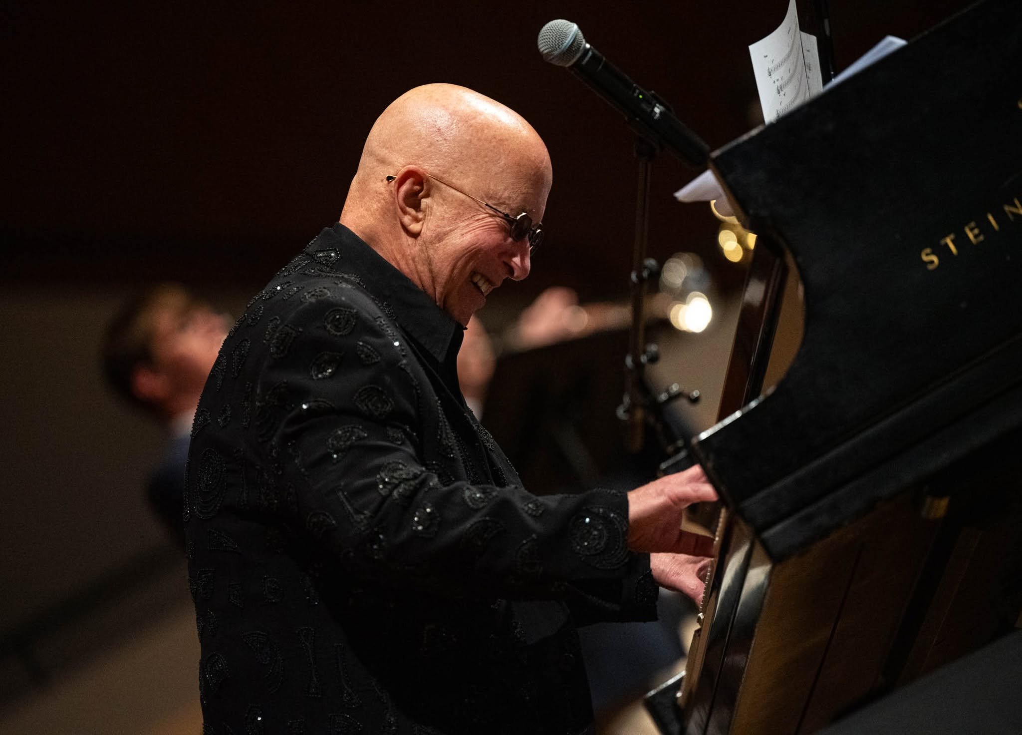 An Paul Shaffer with a bald head and glasses, wearing a black jacket with decorative patterns, playing a grand piano and smiling.