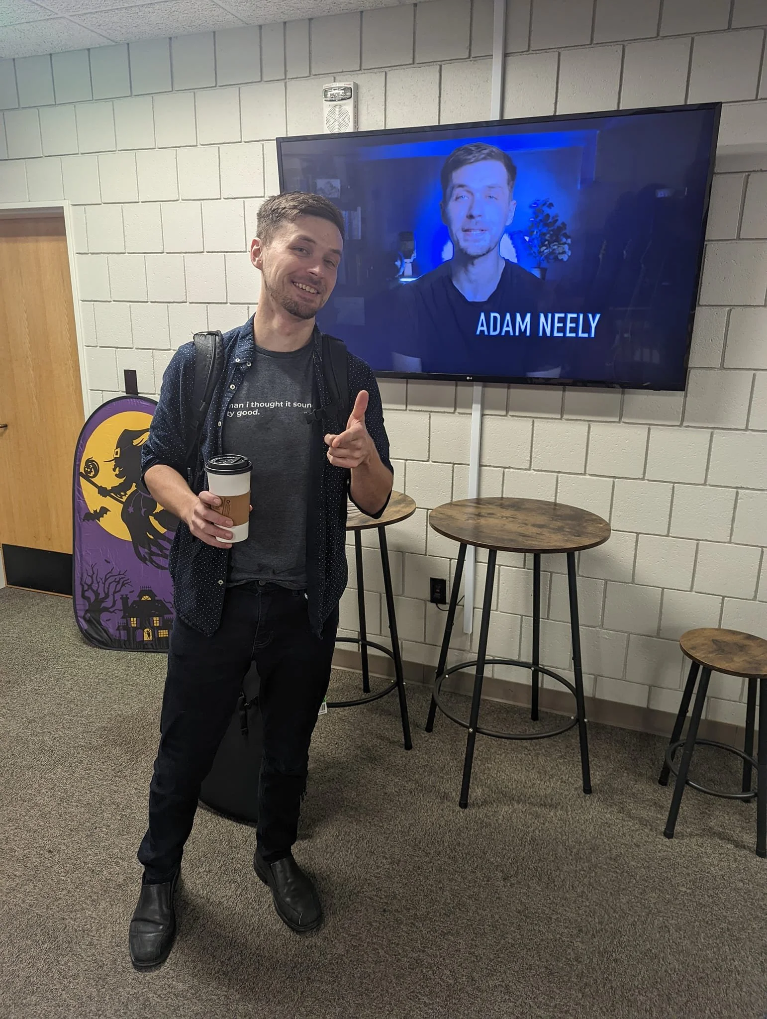 A man named Adam Neely. The man is wearing a dark shirt, a backpack, dark pants, and black boots. Behind him are small round tables and a Halloween-themed bag with a bat and pumpkin design, against a white brick wall.