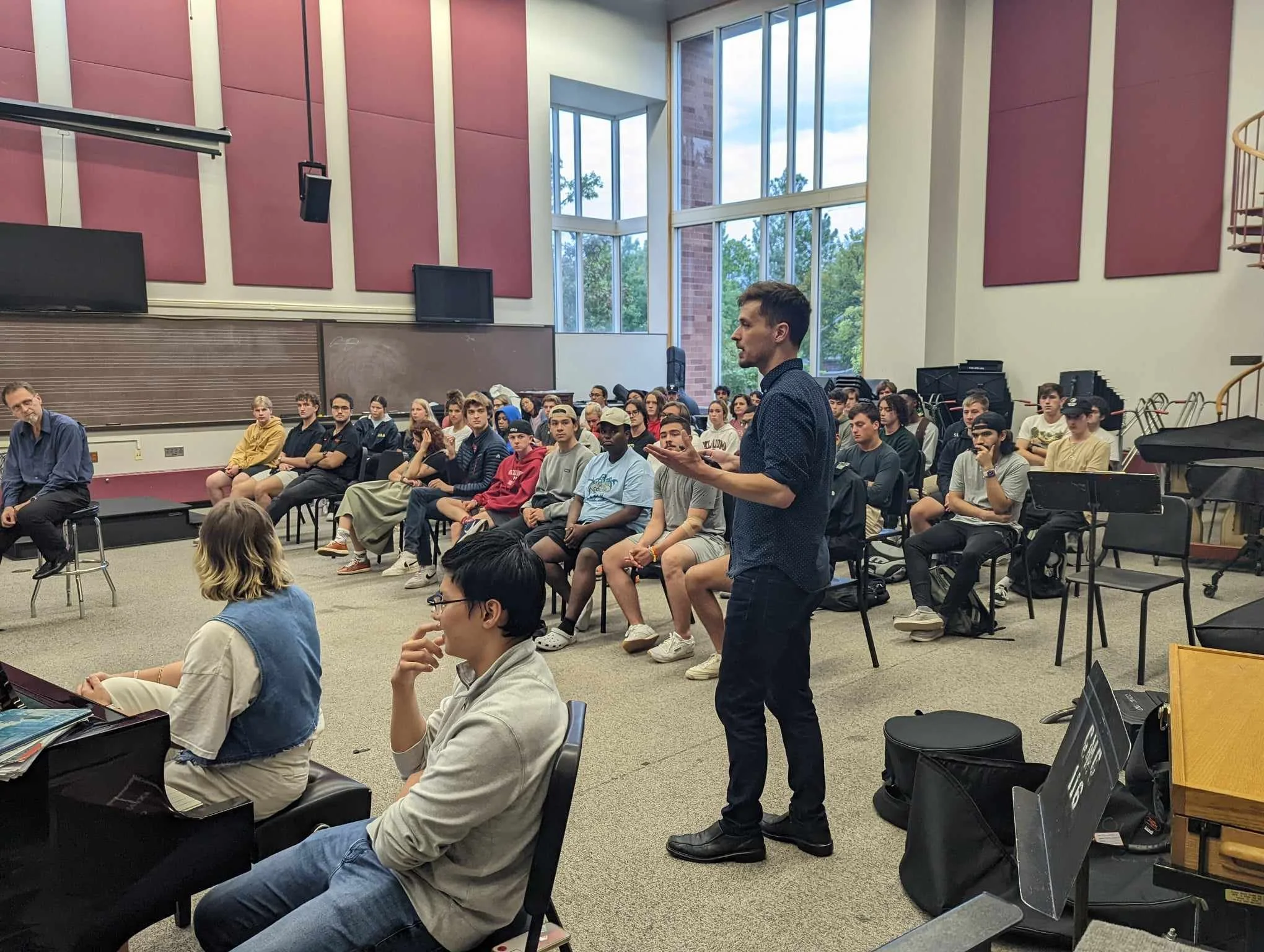 A young man in a dark shirt stands and speaks in front of a classroom filled with students seated in rows, some taking notes, inside a building with large windows and red acoustic panels on the walls.