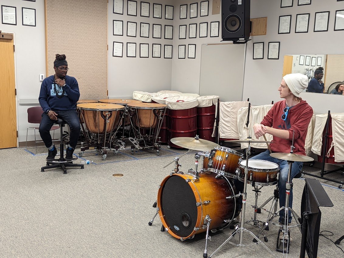 Two musicians practicing with drums in a music room, one on congas and the other on a drum set, with wall certificates and audio equipment in the background.