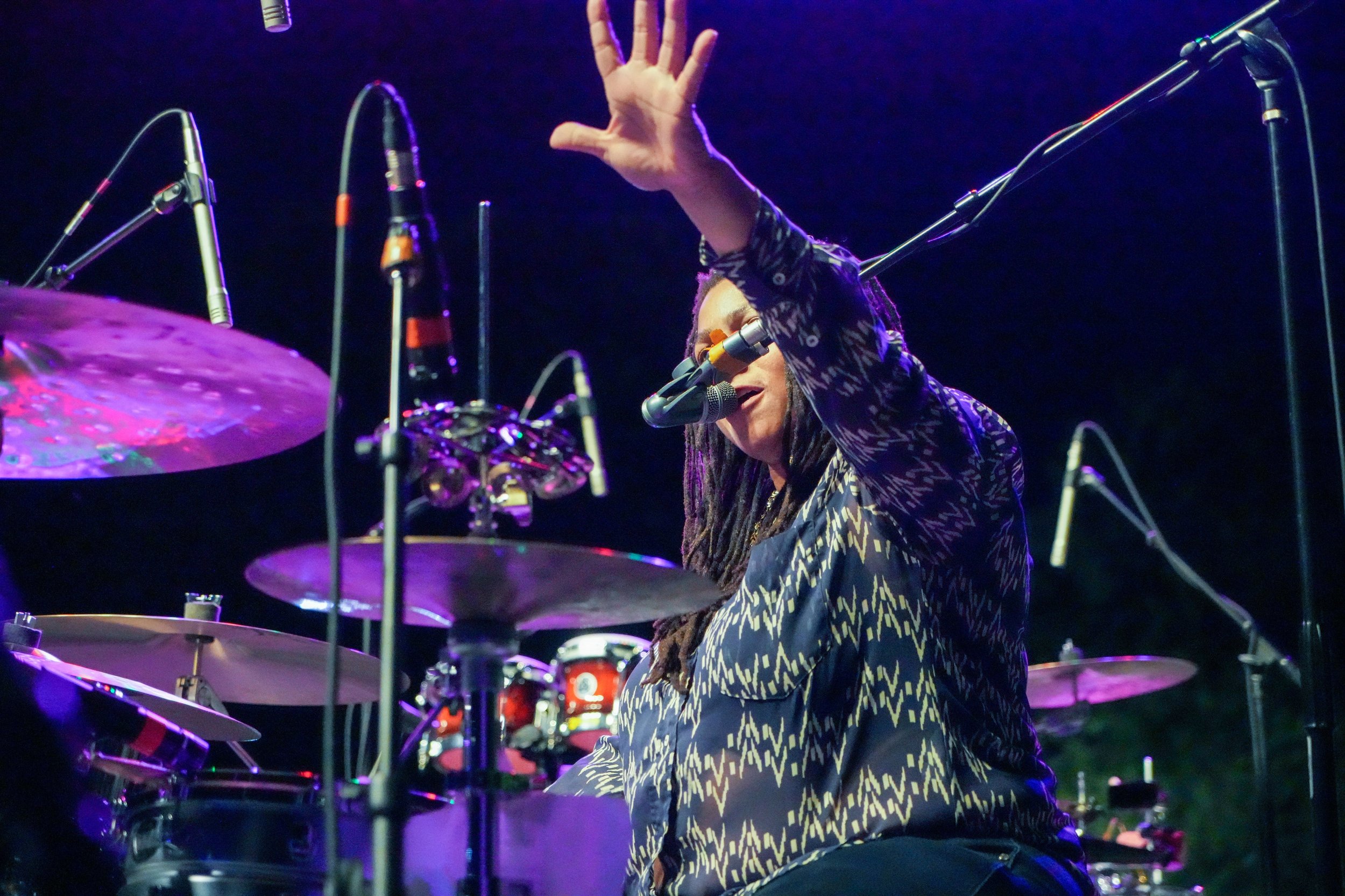 Nikki Glaspie with long hair and glasses, wearing a patterned shirt, is playing drums on a stage with multiple microphones around him. The background is dark, highlighting the stage lighting.