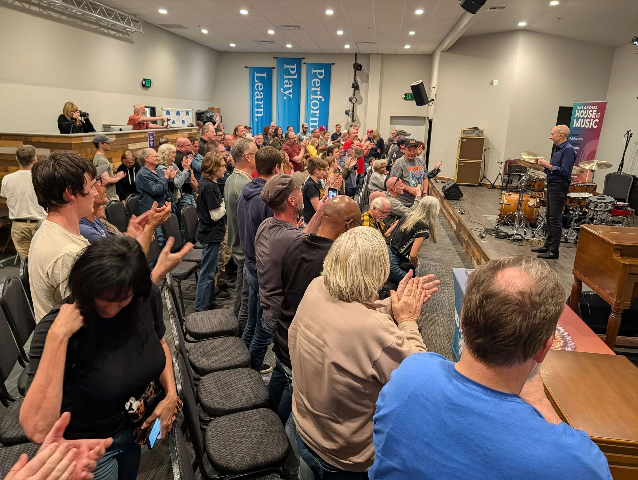 A large audience standing and clapping in a conference room with Steve Smith from Journey on a stage where a musician is holding drumsticks. 