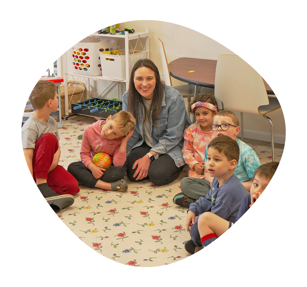 A smiling woman sitting on a colorful carpet with six young children gathered around her in a classroom setting.