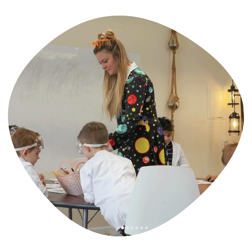 A female teacher in a space-themed jacket supervises children at a table during an activity in a classroom.
