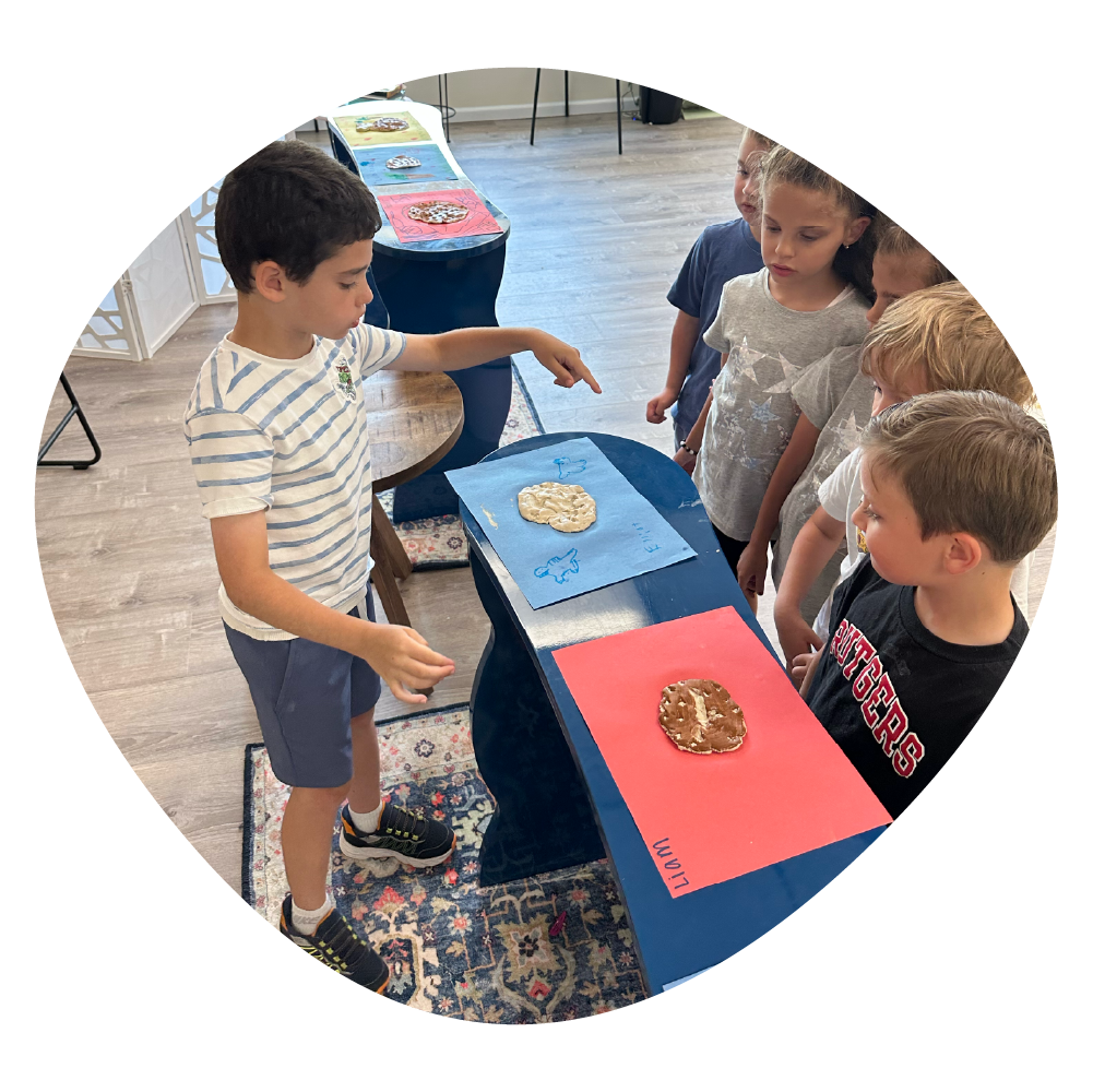 A young boy pointing at two decorated cookies on separate blue and red sheets of paper, as four children watch him intently in a classroom or activity room.