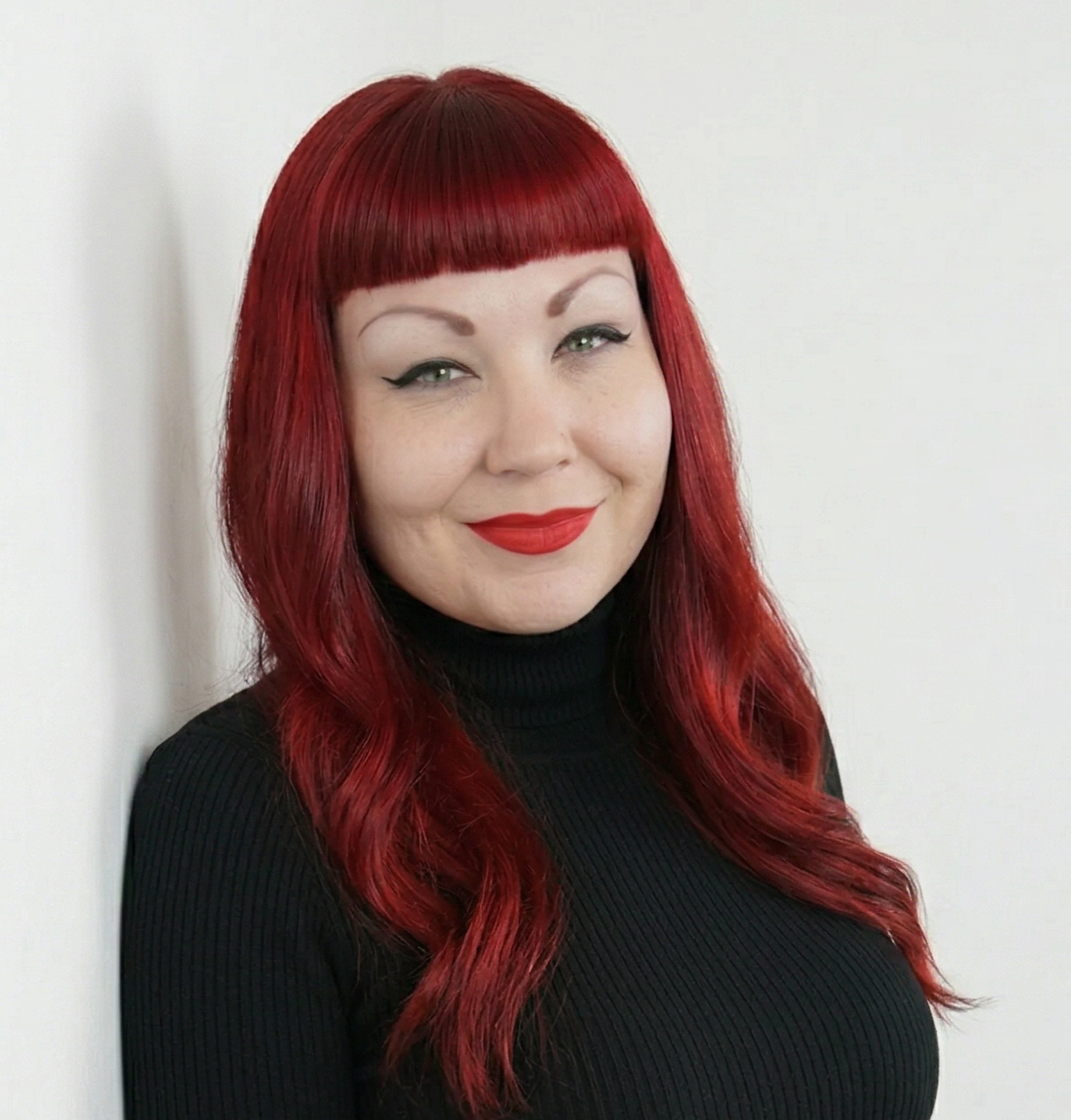 Woman with red hair, red lipstick, black turtleneck, smiling, standing against a plain white wall.