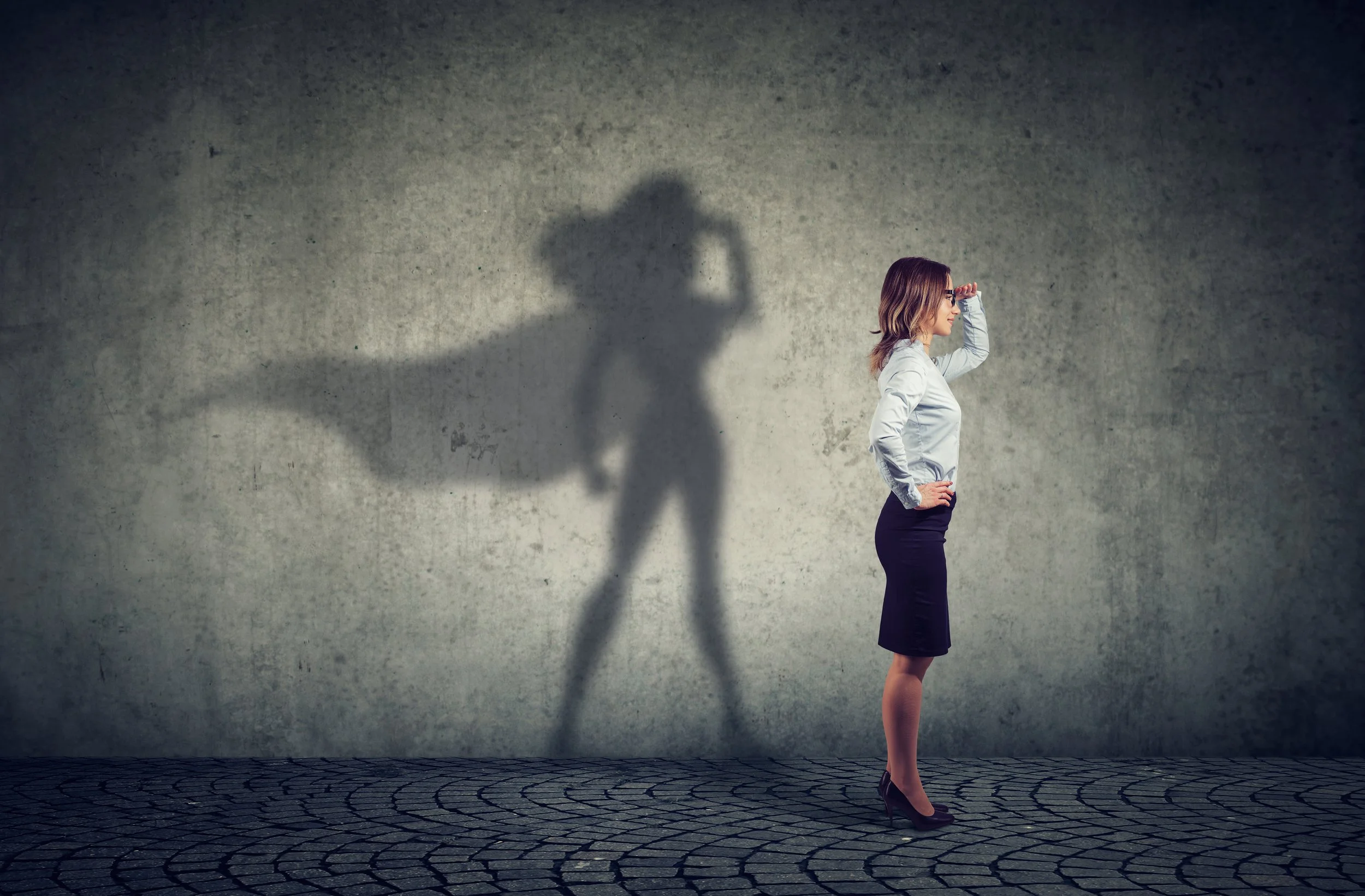 A professional woman stands sideways against a plain wall, with her hand on her forehead, casting a significant shadow that resembles a superhero with a cape.