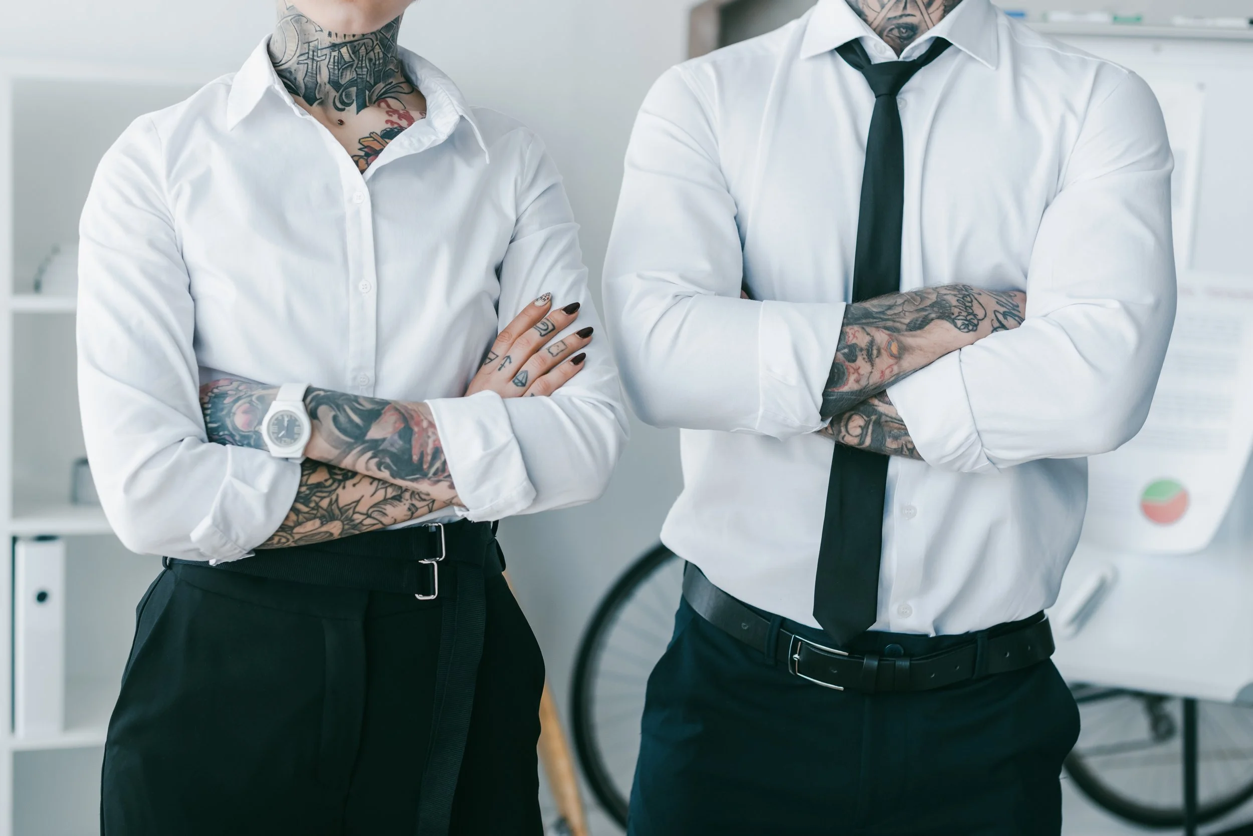 Two people with tattooed arms and necks standing with arms crossed, wearing white shirts and black pants in an office environment.