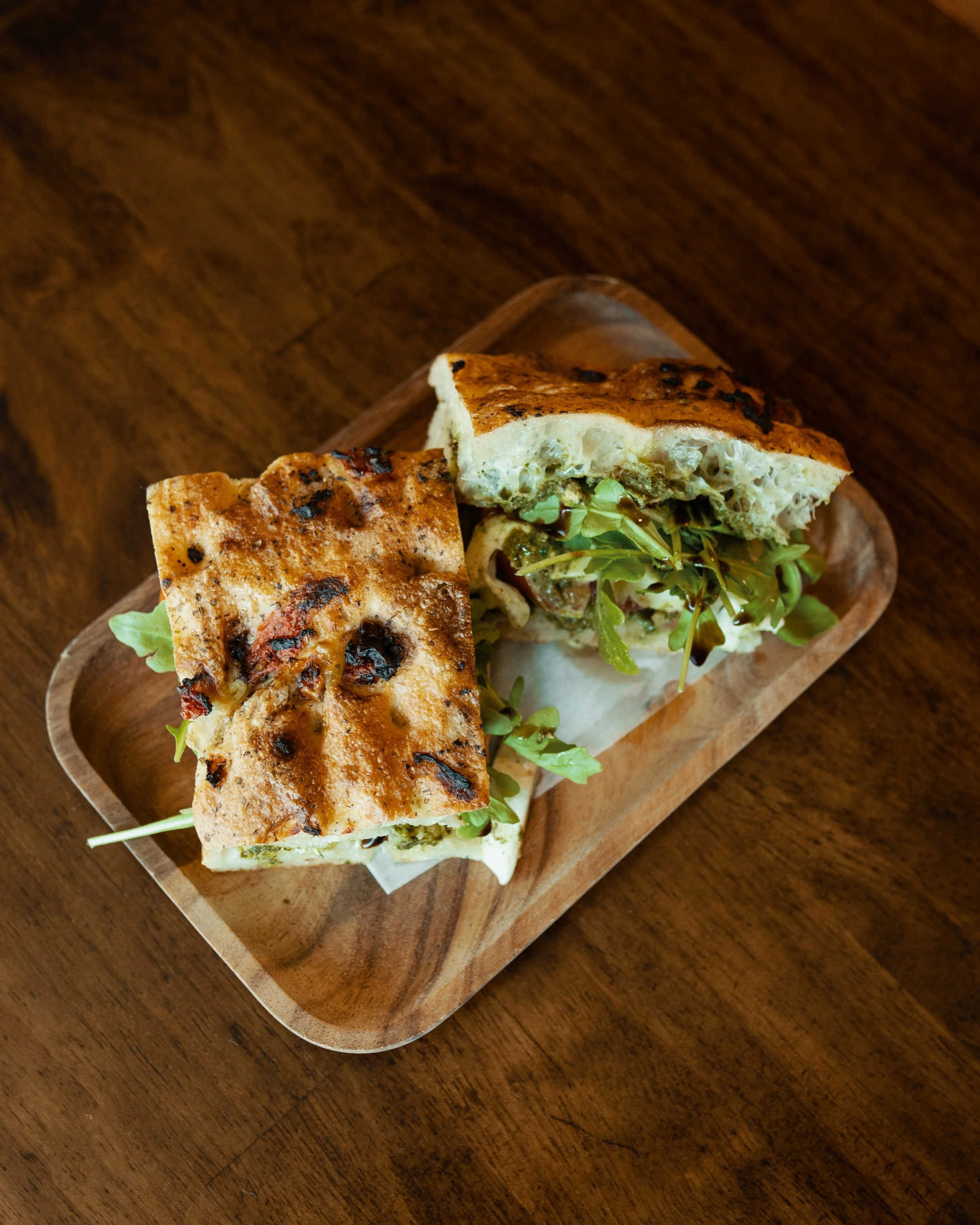 A wooden serving tray with two halves of a sandwich containing lettuce, grilled chicken, and sauce, placed on a wooden table.