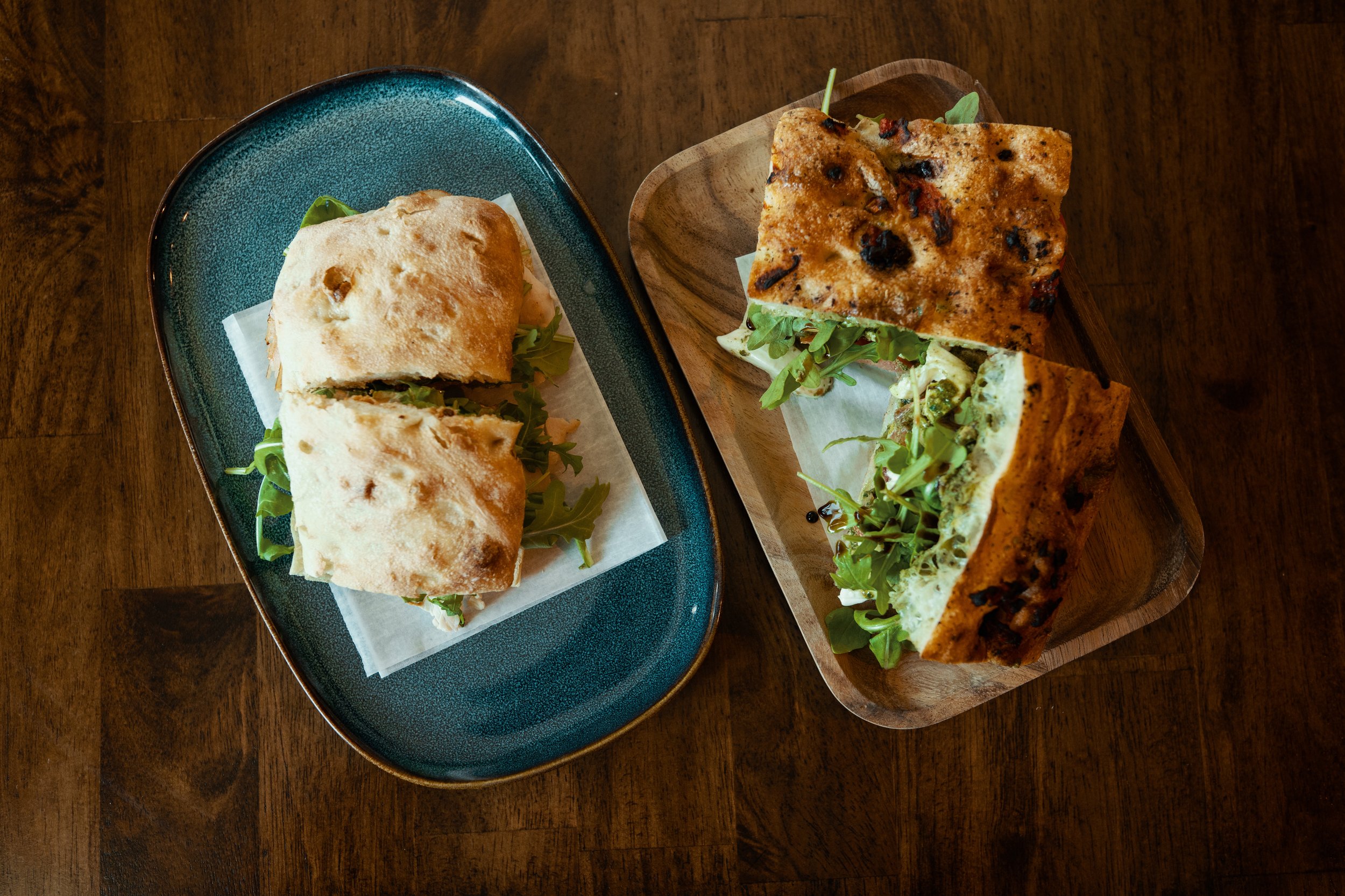 Two sandwiches on wooden and ceramic plates on a wooden table.