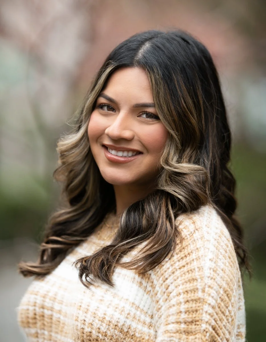 A woman with wavy brown hair with blonde highlights smiling outdoors.
