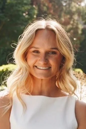 A smiling woman with blonde hair in loose waves, wearing a white top, outdoors with sunlight and trees in the background.