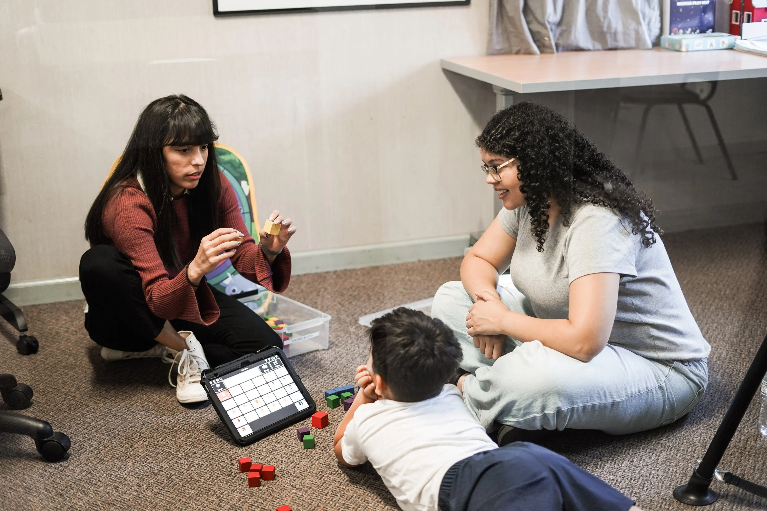 Two women and two children playing a game with colorful cubes and a digital game board on the floor in a room.