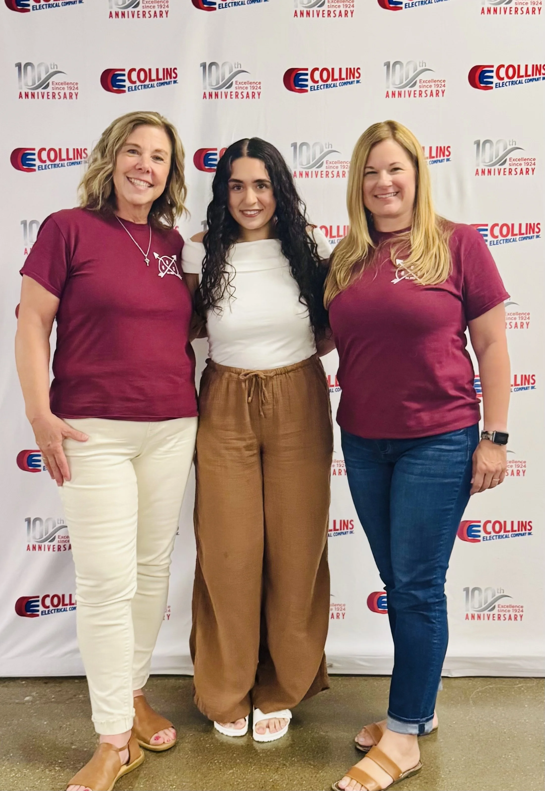 Three women standing together in front of a banner with the logo of Collins Electric and the text celebrating their 100th anniversary. The women are smiling and dressed casually, with two women wearing maroon T-shirts and the woman in the middle wearing a white top and brown pants.