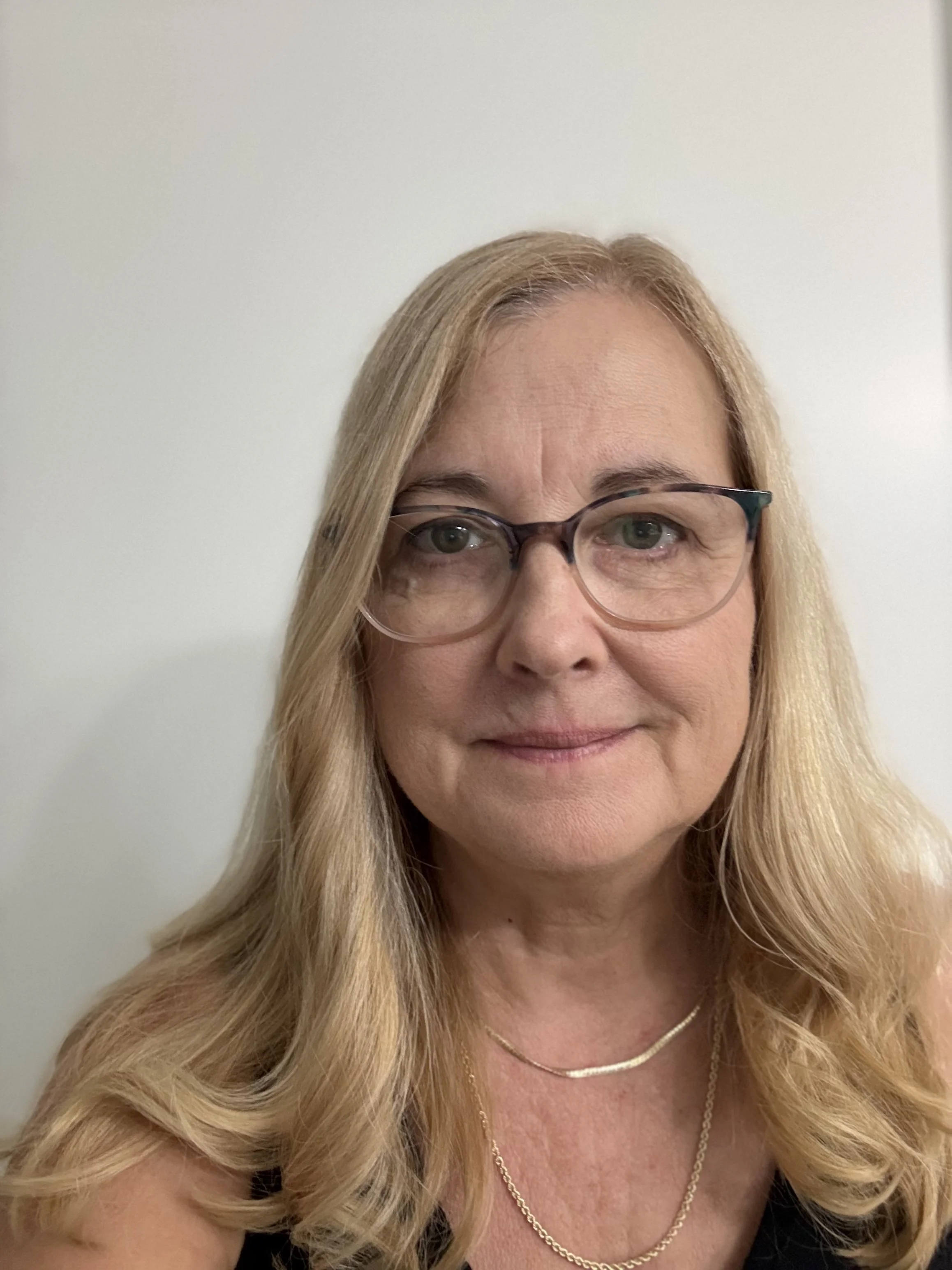 A woman with blonde wavy hair, wearing glasses, a black top, and gold jewelry, smiling against a plain light-colored background.