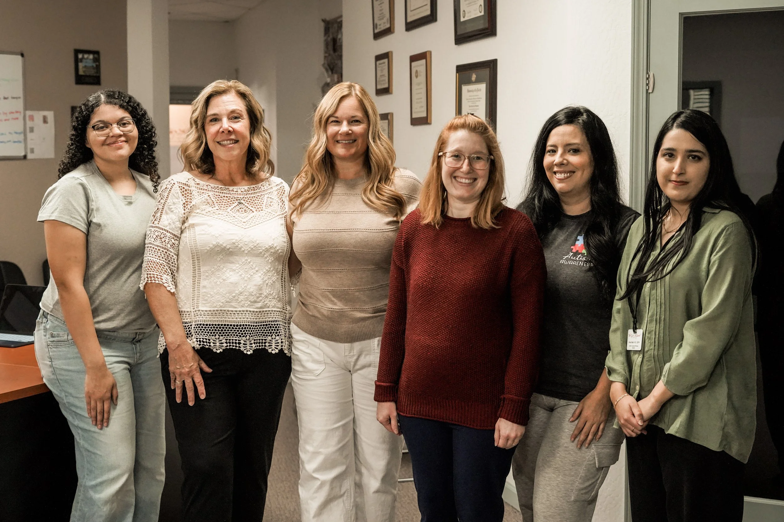Group of six women standing together in an indoor setting, smiling at the camera, with framed certificates on the wall behind them.