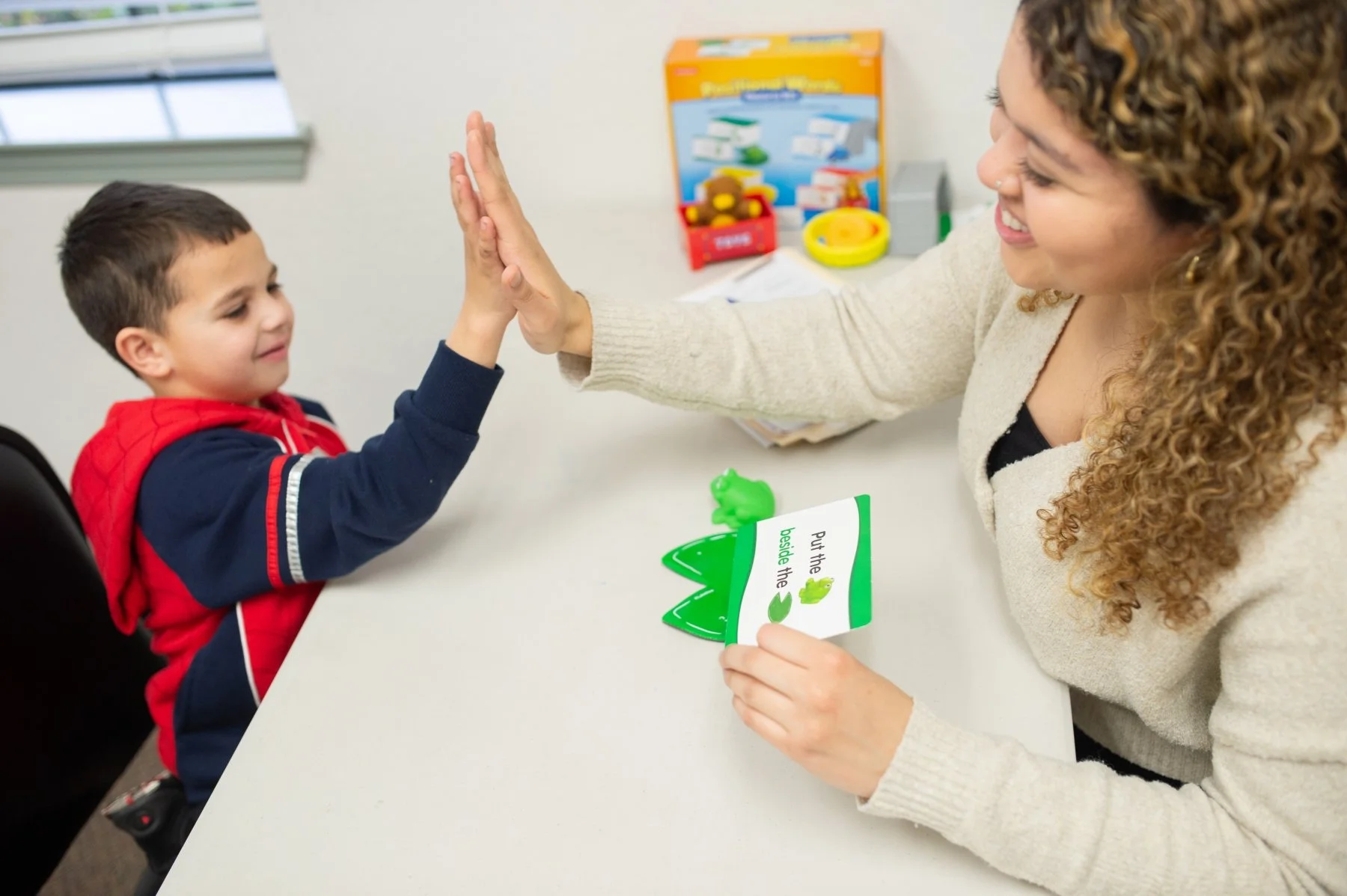 A woman giving a high-five to a young boy sitting at a table during a therapy session. The woman is holding a card with frogs and text, and there are toys and educational items in the background.