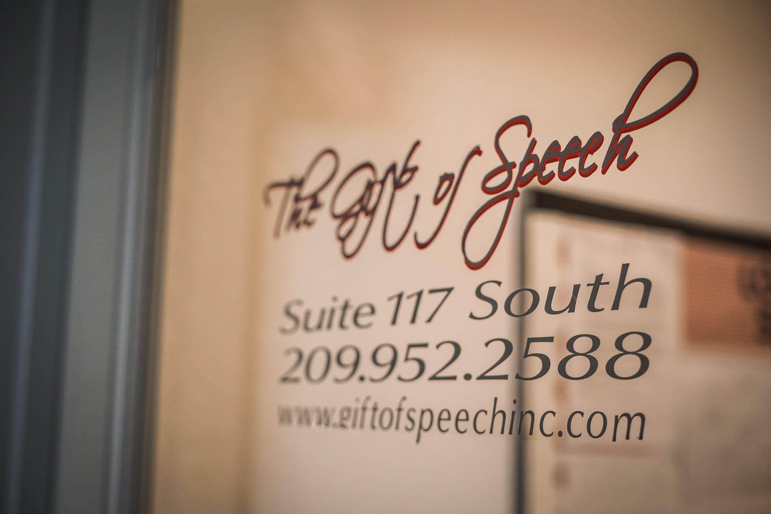 Close-up of a business sign with the text 'The Gift of Speech' in red cursive, and address details including Suite 117 South and a website www.giftofspeechinc.com.