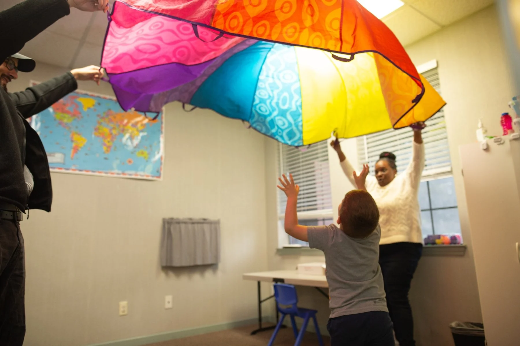 People in a room playing with a colorful rainbow parachute, with a child in front reaching up, a woman holding the parachute, and a man at the side. A world map is on the wall in the background.