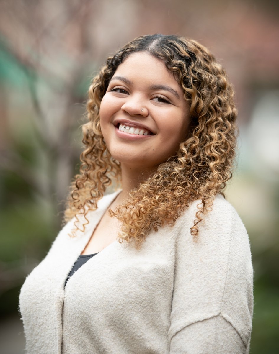 A smiling woman with curly light brown hair, wearing a light beige sweater, standing outdoors with blurred trees in the background.