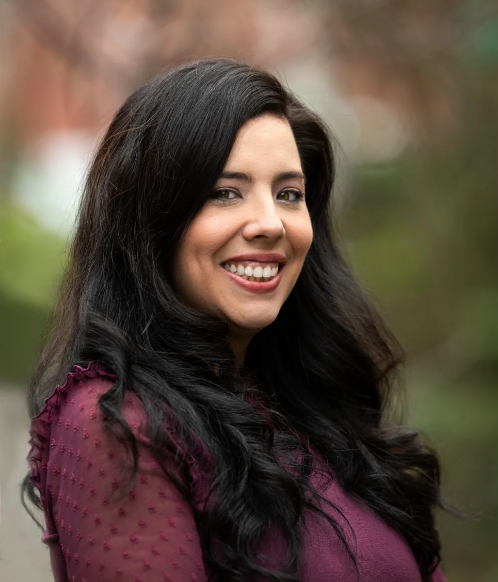 Portrait of a woman with long dark hair smiling outdoors, wearing a purple top with textured sleeves.